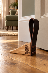 A wooden and leather door stopper resting on a hardwood floor in front of a white door, with a blurred living room interior in the background.
