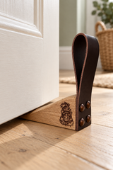 A wooden and leather door stopper resting on a light wood floor in front of a white door, with a small engraved lion logo and three metal rivets visible.