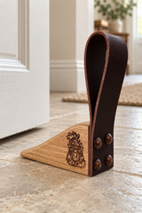 A wooden and leather door stopper placed on a stone floor in front of a white door. The stopper has a triangular wooden base with a dark brown leather loop attached, and a carved animal face design on the front.
