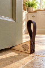 A wooden door stopper with a dark brown leather loop and an engraved tree design, shown wedged between a closed door and a stone floor in a sunlit room.