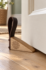 A wooden door stopper with a dark brown leather loop and a triangular wooden base featuring an engraved tree design, shown blocking a white door on a wooden floor.