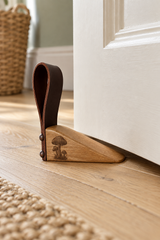 A wooden and leather door stopper with mushroom engraving resting on a light wood floor in front of a white door, with a woven basket and rug in the background.