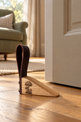 A wooden door stopper with a dark brown leather loop handle and a small mushroom engraving, shown on a wooden floor in front of a white door in a home interior.