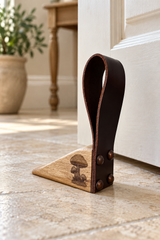 A rustic leather and wood door stopper with mushroom engraving resting on a stone floor in front of a white door, with a blurred indoor background.