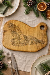 A rustic wooden cutting board with an engraved eagle design, styled on a linen table runner with holiday greenery, dried orange slices, and plates.