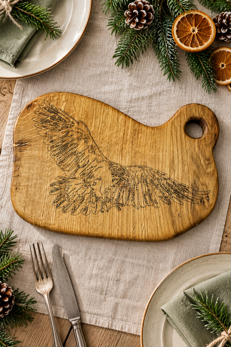 A rustic wooden cutting board with an engraved eagle design, styled on a linen table runner with holiday greenery, dried orange slices, and plates.