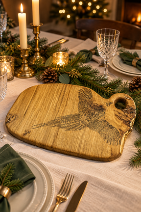 A rustic wooden serving board with a carved pheasant design, styled on a decorated holiday table with candles, pinecones, and plates.
