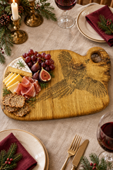 A rustic wooden charcuterie board with a carved pheasant design, styled on a linen tablecloth with cheese, crackers, grapes, and prosciutto, surrounded by plates, wine glasses, candles, and pine branches.