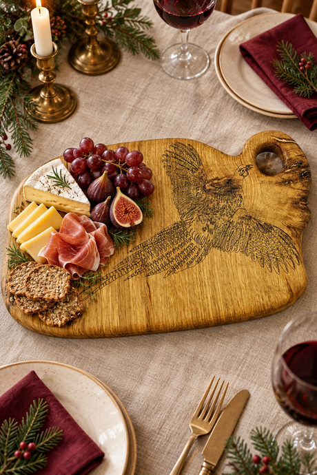 A rustic wooden charcuterie board with a carved pheasant design, styled on a linen tablecloth with cheese, crackers, grapes, and prosciutto, surrounded by plates, wine glasses, candles, and pine branches.