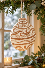 A round terracotta Christmas ornament with brown hand-painted wavy patterns, hanging from a string under pine branches and eucalyptus near a window.