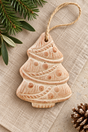 A terracotta Christmas tree ornament in a natural beige stoneware finish with embossed holiday patterns and a hanging twine loop, styled on a textured linen surface with pine branches and a pinecone.