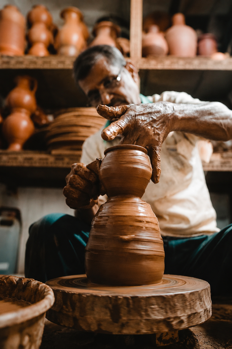 Potter at work shaping a clay pot on a wheel with shelves of finished pots in the background.
