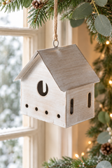 A small house-shaped metal hanging bird house ornament with a weathered light beige finish, hanging from a tree branch with evergreen needles and warm string lights in the background.
