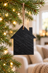 A small black slate house-shaped ornament hanging from a natural twine string on a Christmas tree branch, with warm blurred lights and a cozy living room in the background.