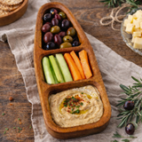 Wooden appetizer tray with hummus, olives, carrots, and celery on a wooden table.