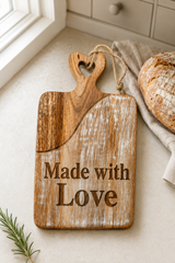 A rustic wooden cutting board with a heart-shaped handle and the engraved words 'Made with Love' in dark brown lettering. The board is styled on a light beige countertop next to a loaf of bread on a cloth napkin, with a window and kitchen tiles in the background.