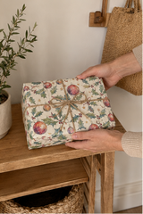 A wrapped rectangular gift box with a vintage holly and bauble print wrapping paper sits on a wooden shelf, held by two hands. The scene is styled with a potted olive branch and a woven jute bag in a warm, neutral indoor setting.
