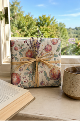 A square gift box wrapped in cream-colored Christmas-themed paper with red ornaments, green holly leaves, and small stars, tied with a natural jute twine bow and decorated with dried lavender sprigs. The box is placed on a rustic wooden windowsill next to an open book and a speckled ceramic mug, with a blurred outdoor garden background visible through the window.
