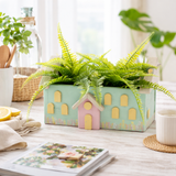 small house-shaped planters with green ferns on a wooden surface.