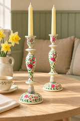 Two decorative wood candlestick holders with cream bases and floral rose patterns, holding white taper candles, set on a light wood table in a cozy living room setting.