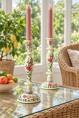 Two floral wood candlestick holders with pink taper candles, displayed on a glass-topped table in a bright room with a wicker chair and potted plant in the background.