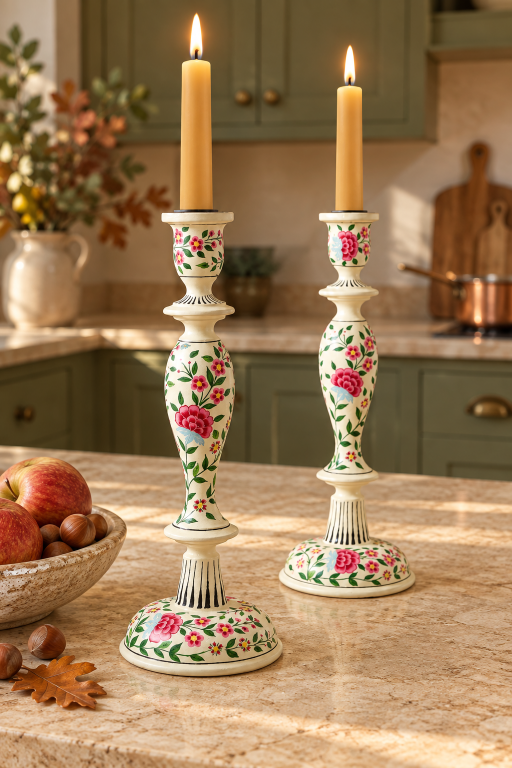 Two ornate ivory coloured wood hand painted candlestick holders with pink and green floral patterns, each holding a lit yellow taper candle, set on a kitchen counter with apples and autumn decor in the background.