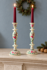 Two ornate ivory coloured wood candlestick holders with pink floral patterns, each holding a lit dark red taper candle, displayed on a stone mantel against a blue-grey wall with a pine wreath in the background.
