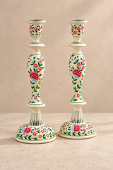 A pair of decorative wood candlesticks with a ivory coloured base and stem, decorated with a vibrant hand-painted floral pattern of pink roses and green leaves, set against a neutral beige background.