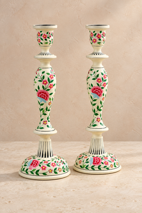 A pair of decorative wood candlesticks with a ivory coloured base and stem, decorated with a vibrant hand-painted floral pattern of pink roses and green leaves, set against a neutral beige background.