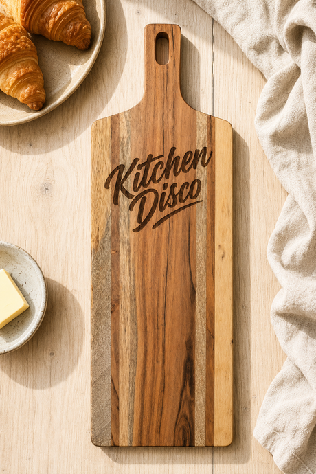 A rectangular wooden serving board with a handle, featuring the words 'Kitchen Disco' engraved in a dark script font. The board is styled in a flat-lay setting on a light wood table, surrounded by food props like croissants, a plate with butter, and a beige linen cloth.