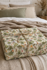 A square gift box wrapped in a Koi Pond -themed wrapping paper with a rustic twine bow, placed on a cozy bed with neutral-toned bedding and pillows.
