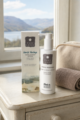 Pure Lakes World Heritage Sweet Basil & Orange Hand & Body Lotion bottle and its matching outer box, styled on a rustic white wooden table next to a beige towel, with a scenic lake and mountains in the background.