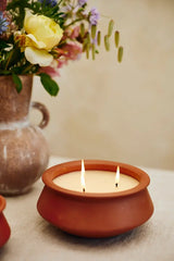 A terracotta clay candle holder with three lit wicks holding a cream-colored candle, set on a light beige surface with a blurred vase of flowers in the background.
Large 3 Wick Terracotta Candle | Lavender & Vanilla