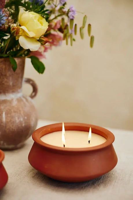 A terracotta clay candle holder with three lit wicks holding a cream-colored candle, set on a light beige surface with a blurred vase of flowers in the background.
Large 3 Wick Terracotta Candle | Lavender & Vanilla