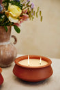 A terracotta clay candle holder with three lit wicks holding a cream-colored candle, set on a light beige surface with a blurred vase of flowers in the background.
Large 3 Wick Terracotta Candle | Lavender & Vanilla