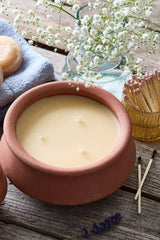 A terracotta clay candle set on a rustic wooden table, featuring a large jar candle and a smaller matching candle. The scene is styled with a blue cloth, white baby's breath flowers, lavender sprigs, and a small gold matchbox.