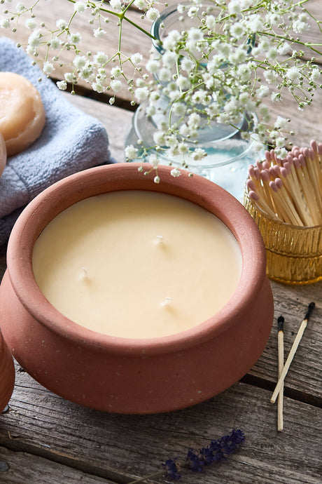 A terracotta clay candle set on a rustic wooden table, featuring a large jar candle and a smaller matching candle. The scene is styled with a blue cloth, white baby's breath flowers, lavender sprigs, and a small gold matchbox.