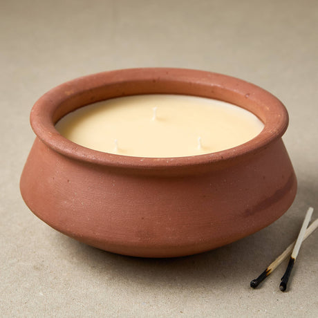 A terracotta clay candle pot filled with creamy off-white wax, photographed on a neutral beige surface with two matches beside it.