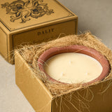 A Dalit brand candle in a square kraft paper box, shown partially open with a natural fiber wrap around the candle vessel. The candle has a matte cream-colored wax surface with three wicks and a dusty rose terracotta-colored rim.