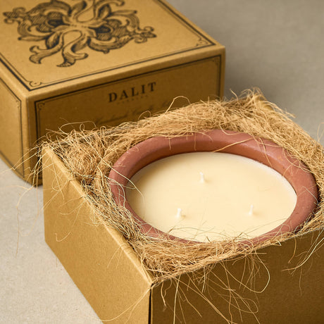 A Dalit brand candle in a square kraft paper box, shown partially open with a natural fiber wrap around the candle vessel. The candle has a matte cream-colored wax surface with three wicks and a dusty rose terracotta-colored rim.