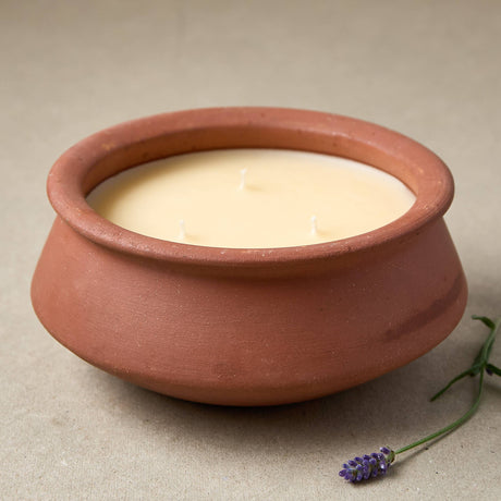 A three-wick soy candle in a terracotta-colored clay pot, styled with a small sprig of lavender on a neutral beige background.