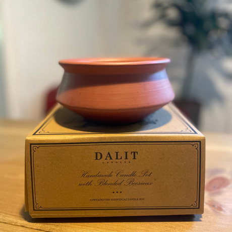 A terracotta-colored ceramic candle holder with a matching lid, displayed on top of its gold-colored Dalit Candle box on a wooden surface.
