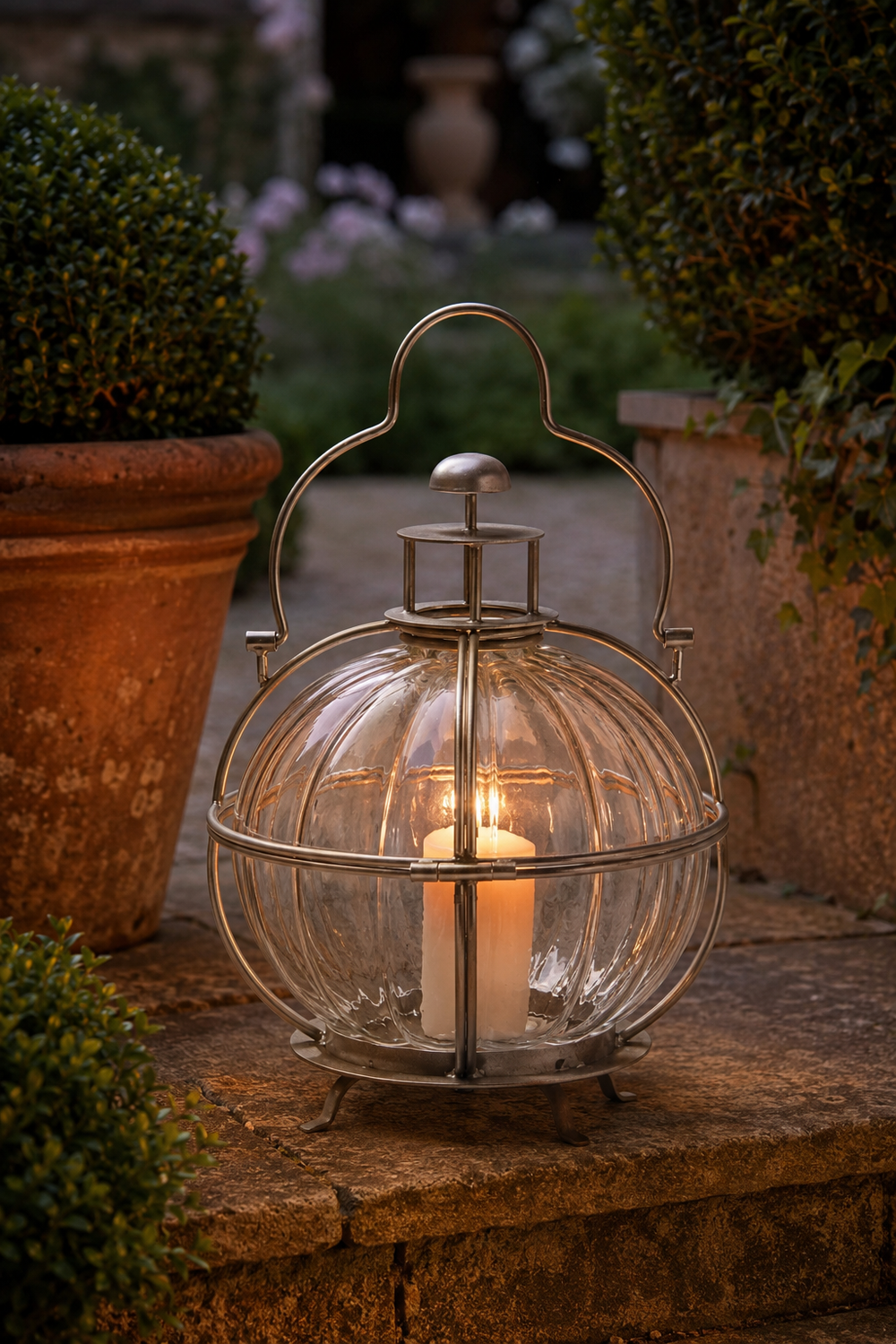 A ribbed clear glass lantern with an antique bronze metal frame and handle, containing a lit white pillar candle, set on a stone garden path between potted plants.