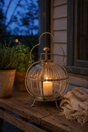 A vintage-style brass lantern with a ribbed glass globe body and a lit pillar candle inside, displayed on a rustic wooden porch table next to potted plants and a throw blanket at dusk.