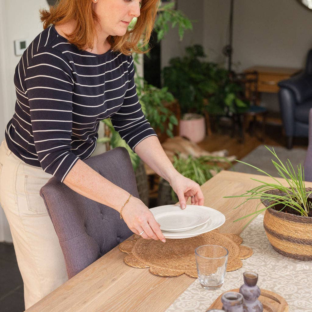 Woman arranging plates on a table with large natural handwoven seagrass placemats with round scalloped edges, in a cozy living room.