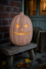 An illuminated terracotta carved pumpkin jack-o'-lantern sits on a rustic wooden bench outdoors against a brick wall and doorway, with fallen leaves on the ground and a throw blanket nearby.