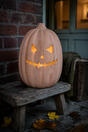 An illuminated terracotta carved pumpkin jack-o'-lantern sits on a rustic wooden bench outdoors against a brick wall and doorway, with fallen leaves on the ground and a throw blanket nearby.