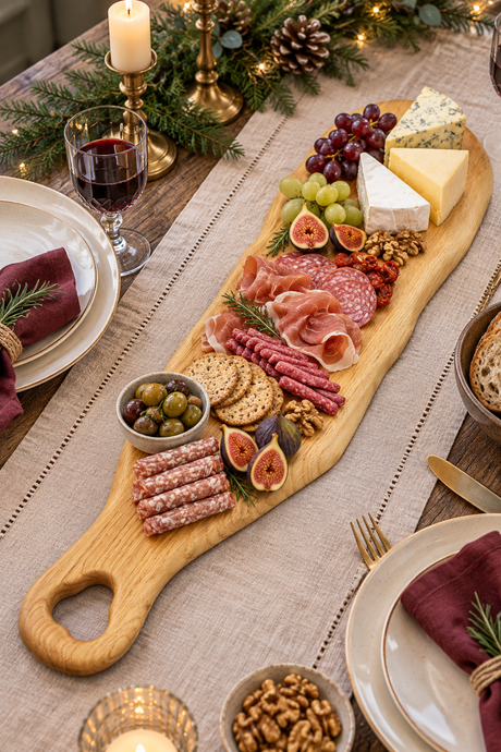 A long wooden charcuterie board arranged with sliced salami, prosciutto, cheese wedges, grapes, figs, crackers, olives, and nuts on a decorated dining table with candles, plates, and greenery.