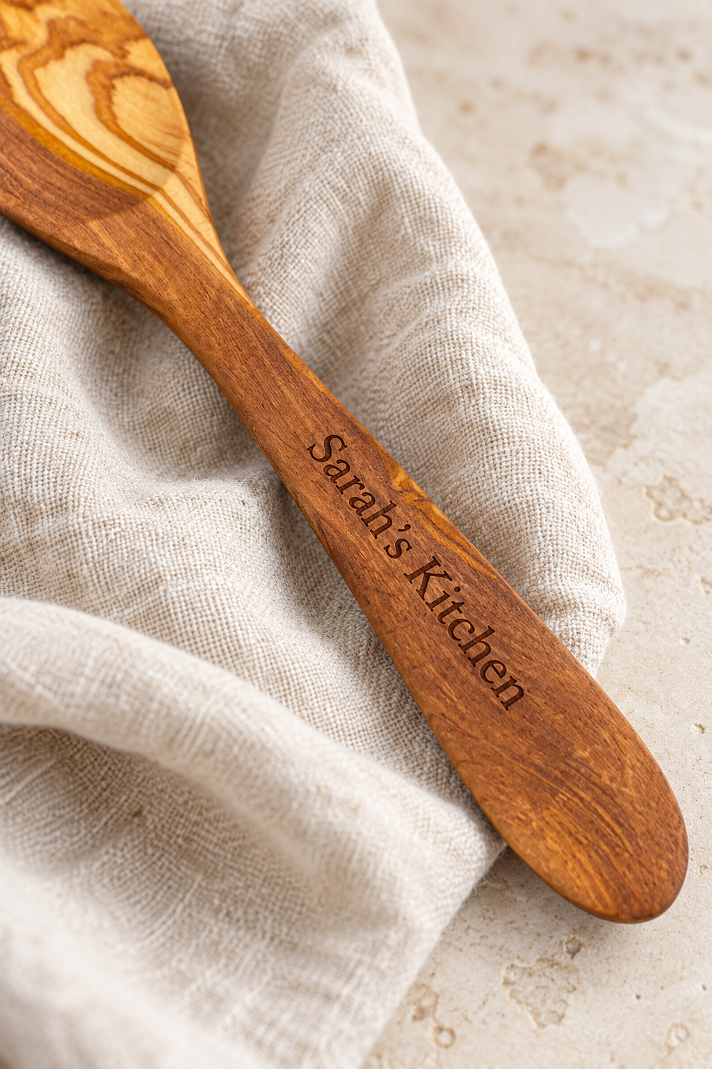Close-up of “Sarah’s Kitchen” engraved into an olive wood spoon handle, showing the fine lettering and natural wood grain against oatmeal linen.