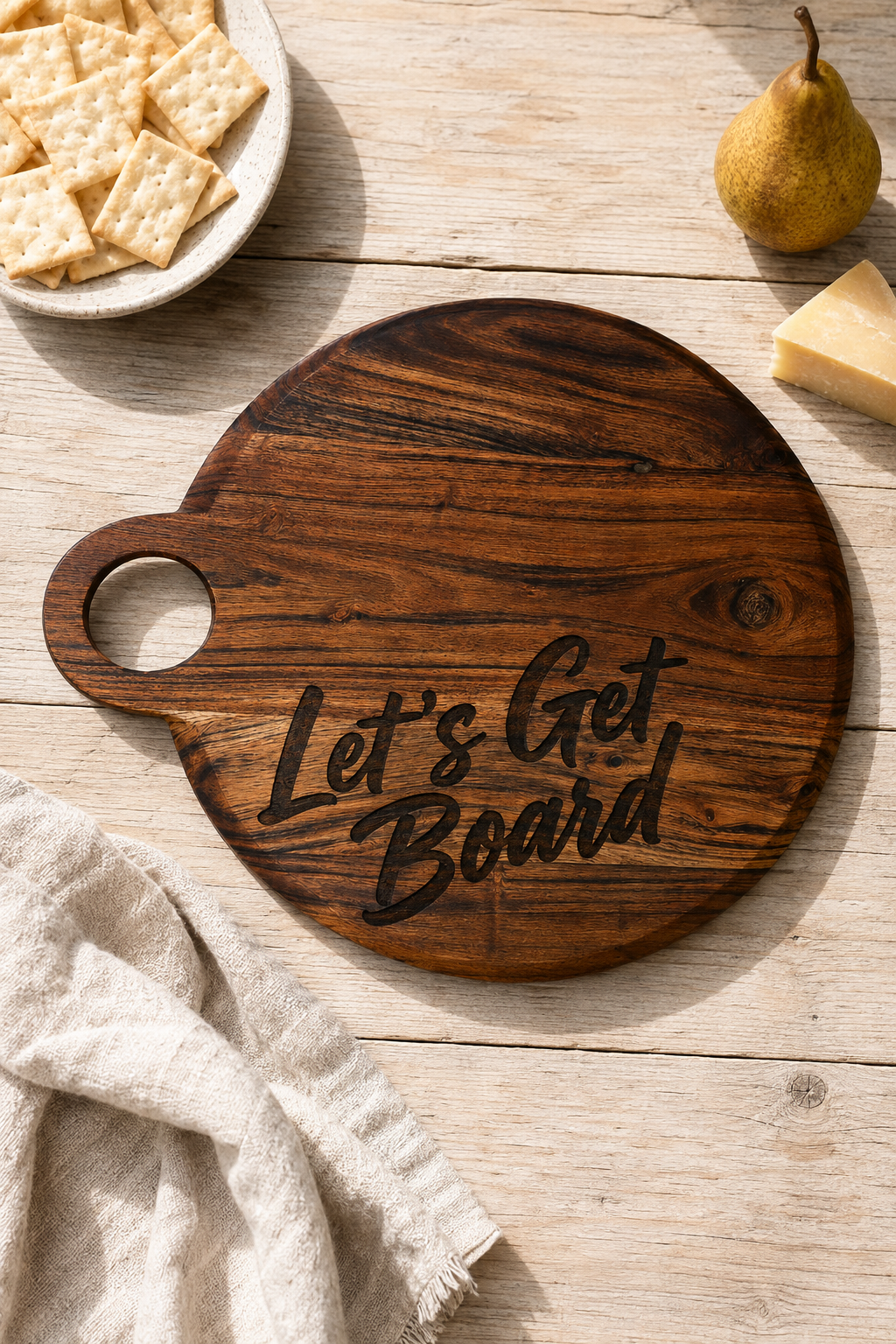 A round dark wood serving board with a handle and the phrase 'Let's Get Board' engraved in black script, styled on a light wood tabletop with crackers, a pear, cheese, and a cloth napkin.